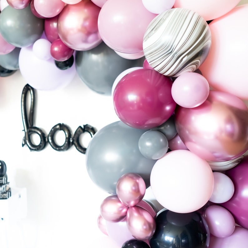 May include: A garland of balloons in shades of pink, gray, and maroon. The balloons are arranged in a circular shape and have a variety of textures, including marble, metallic, and matte.