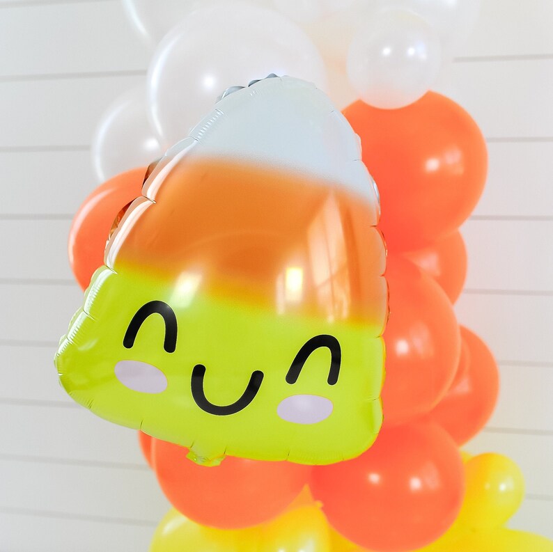 Op de afbeelding: Een gele en oranje ballon in de vorm van een snoepmais met een glimlachend gezicht. De ballon is omringd door oranje, witte en gele ballons.