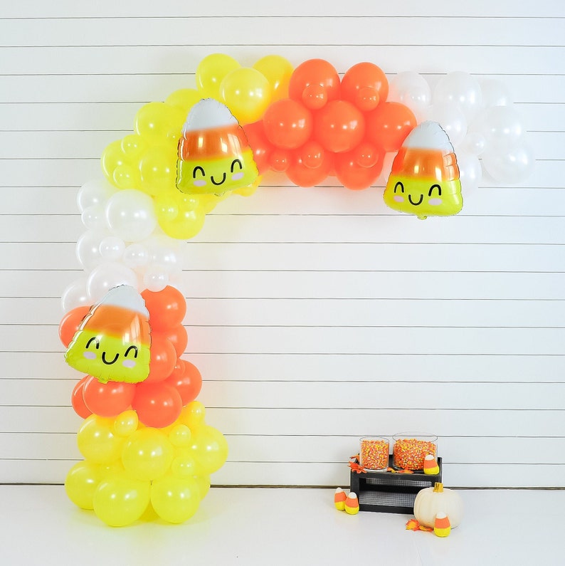 Op de afbeelding: Een witte, oranje en gele ballonboog met snoepmaisontwerpen. De boog is versierd met ballonnen in de vorm van snoepmais met lachende gezichten.