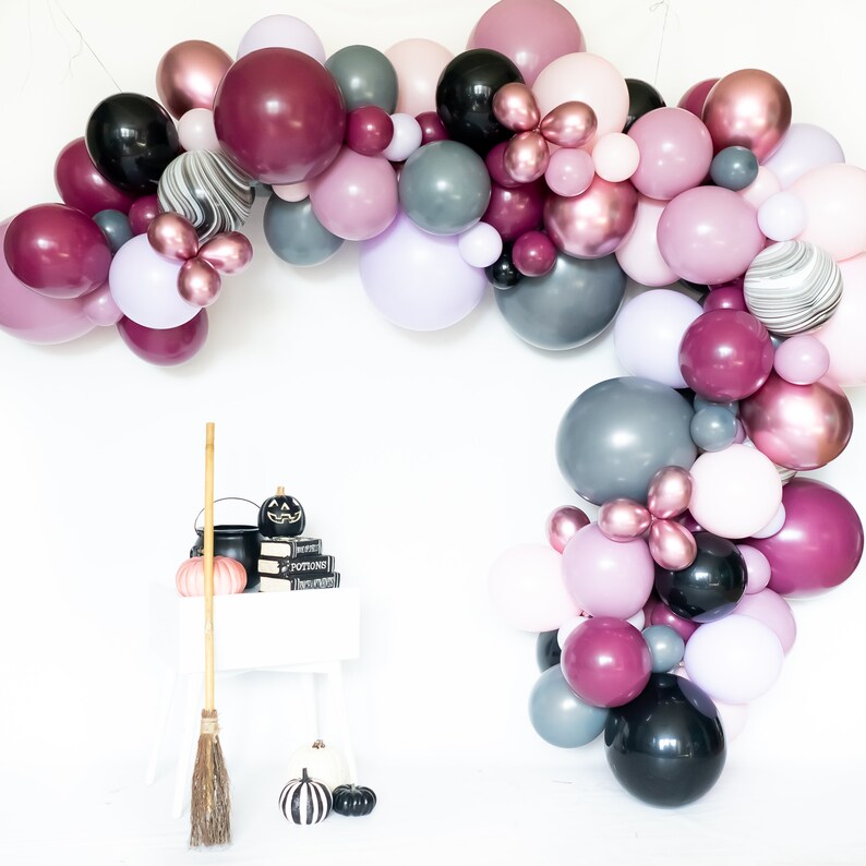 May include: A Halloween party balloon arch with black, burgundy, rose gold, gray, and lavender balloons. The arch is decorated with a black cauldron, books, and a broom.