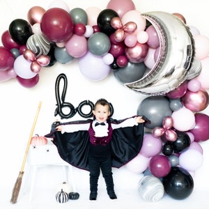 May include: A child dressed as a vampire stands in front of a backdrop of balloons in various shades of pink, purple, gray, and black. The balloons are arranged in an arch shape and include a large silver crescent moon balloon. The child is wearing a black cape, a burgundy vest, a white shirt, and black pants. The child is smiling and holding their arms out to the sides. The word "boo" is spelled out in black balloons in the background.