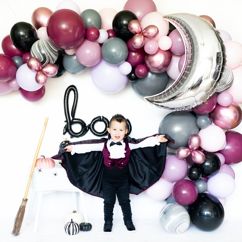 May include: A child dressed as a vampire stands in front of a backdrop of balloons in various shades of pink, purple, gray, and black. The balloons are arranged in an arch shape and include a large silver crescent moon balloon. The child is wearing a black cape, a burgundy vest, a white shirt, and black pants. The child is smiling and holding their arms out to the sides. The word "boo" is spelled out in black balloons in the background.