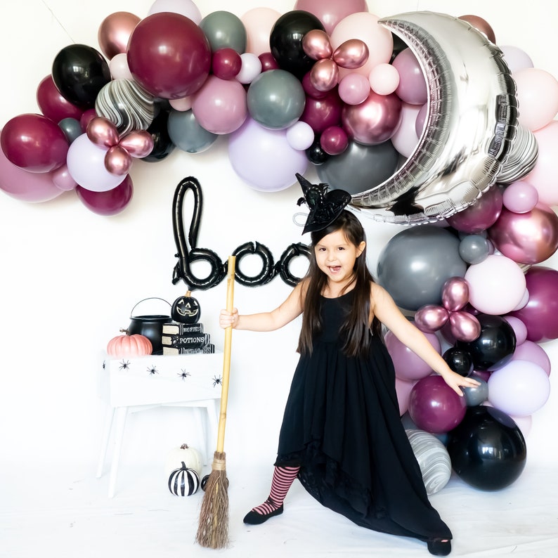 May include: A young girl dressed as a witch holds a broom in front of a black 'boo' sign and a backdrop of purple, pink, and silver balloons. She is wearing a black dress, a witch hat, and striped tights.