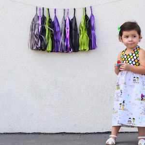 May include: A young girl wearing a white dress with a black and yellow polka dot bodice and a Halloween-themed print. The dress has a gathered skirt and is sleeveless. The girl is standing in front of a white wall with a string of colorful tissue paper tassels hanging above her.