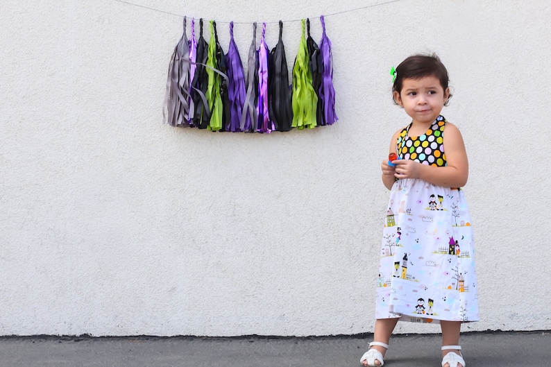 May include: A young girl wearing a white dress with a black and yellow polka dot bodice and a Halloween-themed print. The dress has a gathered skirt and is sleeveless. The girl is standing in front of a white wall with a string of colorful tissue paper tassels hanging above her.
