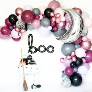May include: A Halloween balloon arch with a silver moon balloon, black and white marble balloons, burgundy, pink, and gray balloons. The arch is decorated with a black "boo" banner and a black cauldron with a witch's broom.