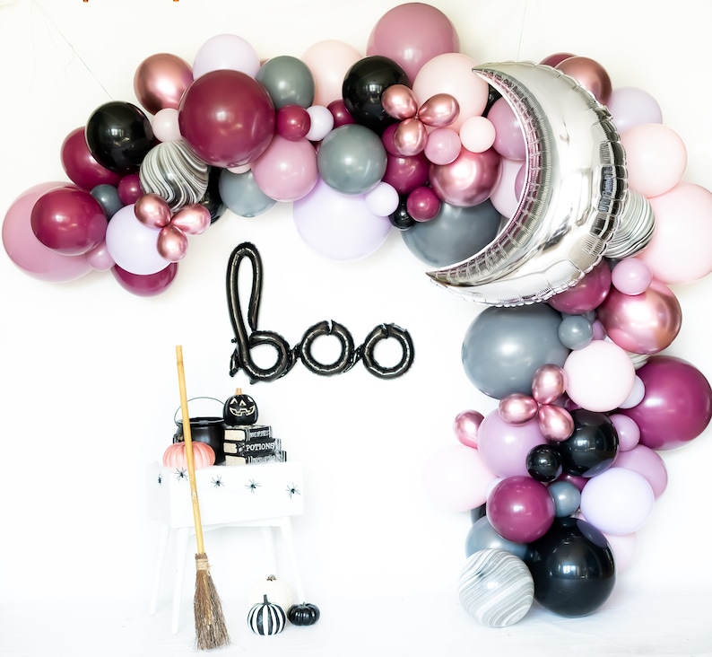 May include: A Halloween balloon arch with a silver moon balloon, black and white marble balloons, burgundy, pink, and gray balloons. The arch is decorated with a black "boo" banner and a black cauldron with a witch's broom.