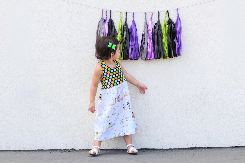 May include: A young girl wearing a black and white polka dot sleeveless dress with a white skirt featuring a whimsical print of houses and figures. She is standing in front of a white wall with a colorful garland of tissue paper tassels hanging above her.