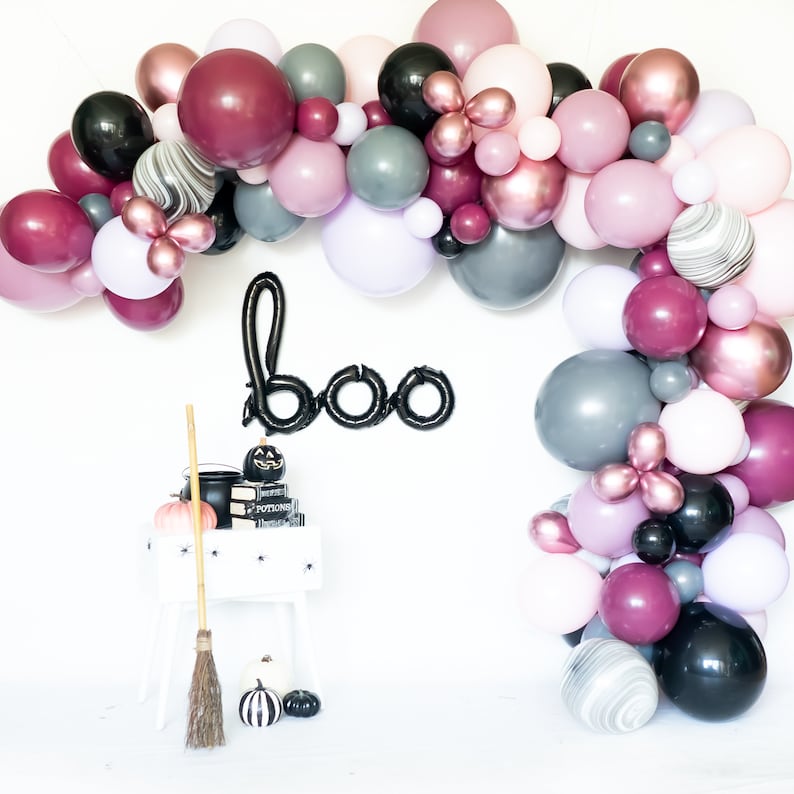 May include: A Halloween balloon arch with black, burgundy, pink, and gray balloons. The arch is decorated with the word "boo" in black balloons. There is a black cauldron and a broom in the foreground.