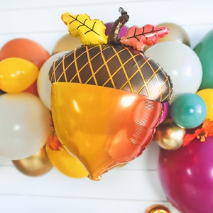 May include: A golden brown and orange acorn shaped balloon with brown leaves and a brown stem. The balloon is surrounded by a variety of colorful balloons, including teal, yellow, white, and pink.