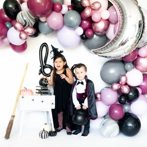 May include: Two children dressed in Halloween costumes stand in front of a wall decorated with a balloon arch. The girl is wearing a black dress and a witch hat. The boy is wearing a black cape and a red velvet vest. The balloon arch is made of purple, pink, and gray balloons. There are also some silver and black balloons in the arch.