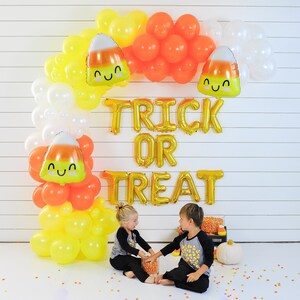 Op de afbeelding: Twee kinderen staan voor een ballonnenboog versierd met oranje, gele en witte ballonnen in de vorm van ma&iuml;s. De ballonnen spellen "Trick or Treat" in gouden letters.