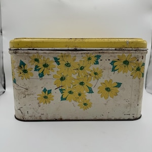 May include: Vintage rectangular metal tin with a yellow lid and white body. The tin is decorated with yellow daisy flowers and teal green leaves. The tin shows signs of wear and tear, adding to its vintage charm.