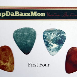 May include: Four guitar picks with different designs. The picks feature a blue and white marble pattern, a blue and white galaxy pattern, a red and orange marble pattern, and a red and orange galaxy pattern. The text "SlapDaBassMon Custom Guitar Picks" is visible at the top of the image. The text "First Four" is visible at the bottom of the image.