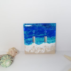May include: A decorative double light switch cover with a beach-themed design. The top portion features shades of blue and turquoise, resembling ocean waves. The bottom has a sandy beige color, with white accents. The cover has two switch openings.