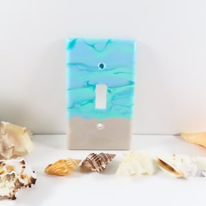 May include: A light switch cover with a blue and white marbled design, resembling ocean waves. The bottom half of the cover is a light beige color, representing sand.