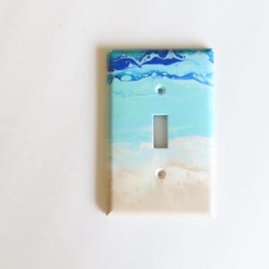 May include: A decorative light switch cover with a beach-themed design. The top portion features a dark blue and white wave pattern, transitioning to light blue and sandy beige at the bottom. The cover has a single toggle switch opening and two screw holes.