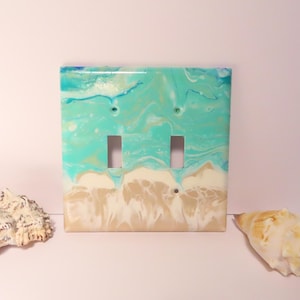 May include: A double light switch cover with a beach-themed design. The cover features a turquoise and white marbled pattern resembling ocean waves and a sandy beach. Two seashells are placed on either side of the switch plate.