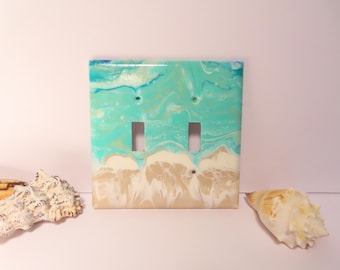 May include: A double light switch cover with a beach-themed design. The cover features a turquoise and white marbled pattern resembling ocean waves and a sandy beach. Two seashells are placed on either side of the switch plate.