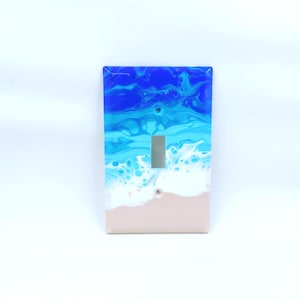 May include: A light switch cover with an abstract ocean scene. The cover features blue, white, and light brown paint, resembling waves crashing on a sandy beach.