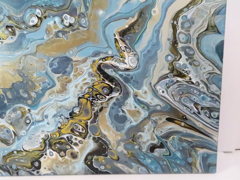 May include: Abstract acrylic pour painting with a fluid design. The artwork features a mix of colors, including shades of blue, gold, and white, creating a marbled effect. The painting has a glossy finish and a unique, organic pattern.
