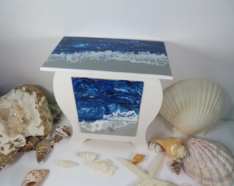 May include: White wooden jewelry box with a blue and silver ocean wave design. The box has a drawer and is decorated with seashells.