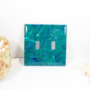 May include: A teal and blue abstract light switch cover with two toggle switches.