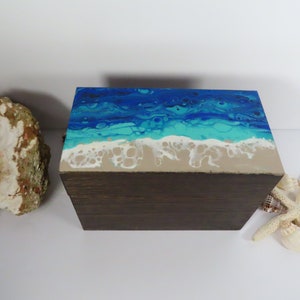 May include: A wooden box with a painted ocean scene on the top. The scene features blue and turquoise water with white foam waves crashing on a sandy beach.