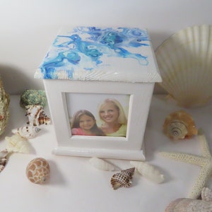May include: White wooden photo box with a blue and white abstract painted lid. The box has a framed opening with a photo of two people. Seashells and a starfish surround the box.