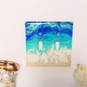 May include: A decorative double light switch cover with a beach-themed design. The cover features a resin design with shades of blue, turquoise, and white, resembling ocean waves and sand. Two small, clear, round screws are visible.
