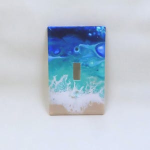 May include: A decorative light switch cover featuring an ocean-themed design. The cover has a rectangular shape with a silver switch. The artwork depicts a beach scene with shades of blue, turquoise, white, and tan.