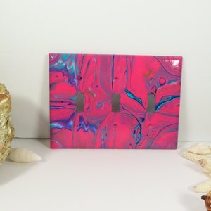 May include: A pink, blue, and white abstract painted light switch cover with three toggle switches.