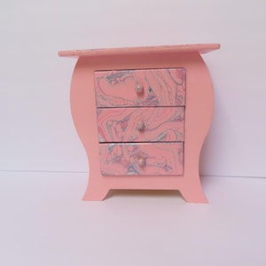 May include: A small pink wooden dresser with three drawers. The drawers have a marbled design in pink, blue, and white.