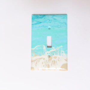 May include: A decorative light switch cover with a beach-themed design. The cover features a turquoise and white wave pattern, with a white toggle switch in the center. The cover is rectangular and has two screw holes.