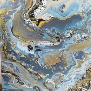 May include: Abstract acrylic pour painting with a fluid design. The artwork features a mix of colors, including blues, gold, white, and gray, creating a marbled effect. The painting has a glossy finish and is stretched on a canvas.