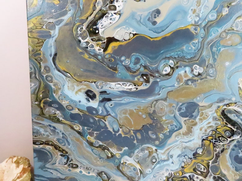 May include: Abstract acrylic pour painting with a fluid design. The artwork features a mix of colors, including blues, gold, white, and gray, creating a marbled effect. The painting has a glossy finish and is stretched on a canvas.