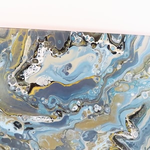 May include: Abstract acrylic painting with a fluid design. The artwork features swirling patterns in shades of blue, gold, gray, and white. The paint appears to have been poured and swirled to create a marbled effect, suitable for wall decor.
