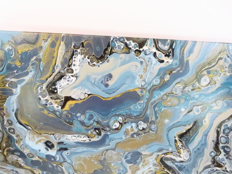May include: Abstract acrylic painting with a fluid design. The artwork features swirling patterns in shades of blue, gold, gray, and white. The paint appears to have been poured and swirled to create a marbled effect, suitable for wall decor.