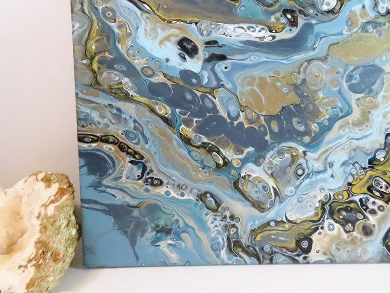 May include: Abstract acrylic painting on canvas with a fluid design. The artwork features a blend of blues, gold, and gray hues, creating a marbled effect. The painting's texture and color palette evoke a sense of depth and movement, suitable for home decor.