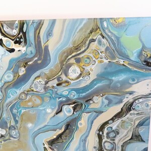 May include: Abstract acrylic painting on canvas featuring flowing patterns in shades of blue, gray, gold, and white. The artwork has a fluid, marbled appearance, with a glossy finish. The canvas is stretched and ready to hang.