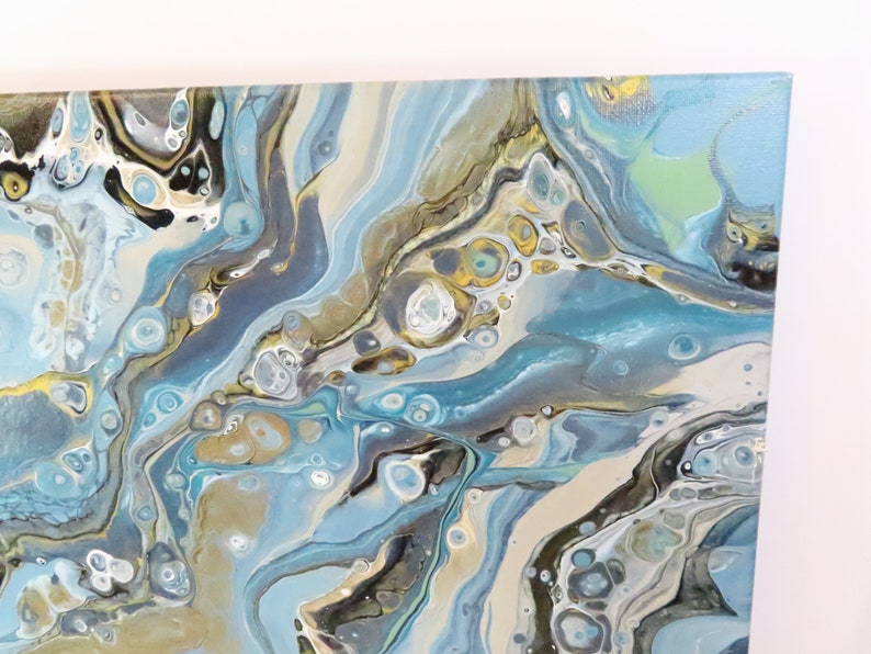 May include: Abstract acrylic painting on canvas featuring flowing patterns in shades of blue, gray, gold, and white. The artwork has a fluid, marbled appearance, with a glossy finish. The canvas is stretched and ready to hang.