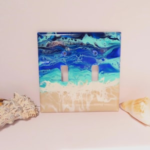 May include: A double light switch cover with an ocean-themed design. The cover features a layered effect with shades of blue, turquoise, and white, resembling waves and a sandy beach. Two seashells are placed on either side of the switch plate.