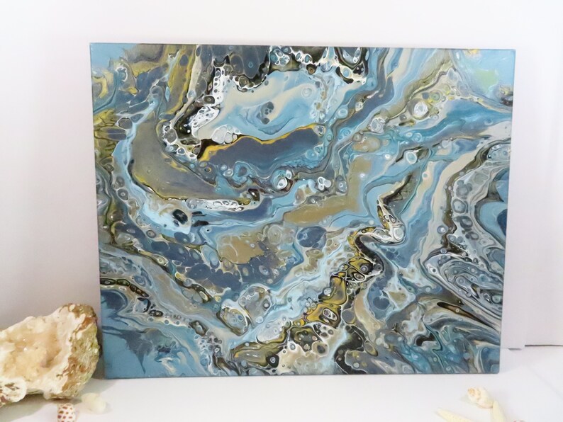 May include: Abstract acrylic pour painting on canvas. The artwork features a fluid design with shades of blue, gray, gold, and white. The colors blend and swirl, creating a marbled effect. The painting is a square format.