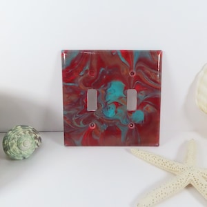 May include: A red and teal abstract swirl design light switch cover with two toggle switches.
