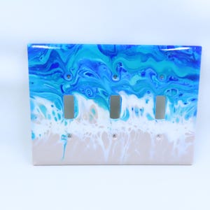 May include: A light switch cover with an abstract ocean scene painted in shades of blue, white, and beige. The cover has three switch openings.