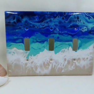 May include: A decorative triple light switch cover with an ocean-themed design. The cover features a glossy finish with layers of blue, turquoise, and white resin, resembling waves and a sandy beach. The cover has three switch openings.
