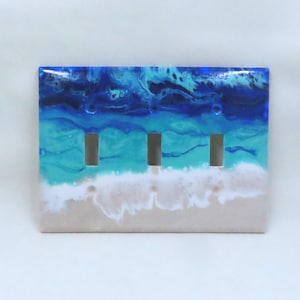 May include: A decorative triple light switch cover with a beach-themed design. The cover features a gradient of colors, including shades of blue, turquoise, white, and tan, resembling an ocean scene. The cover has three rectangular cutouts for the light switches.