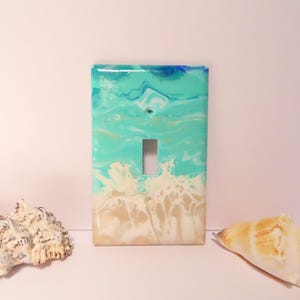 May include: A decorative light switch cover with a beach-themed design. The cover features a turquoise, white, and beige abstract pattern resembling ocean waves and sand. Two seashells are placed on either side of the switch cover.