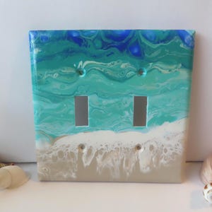 May include: A double light switch wall plate featuring an acrylic pour design that mimics an ocean shoreline. The surface displays swirling shades of deep blue, turquoise, and teal transitioning into white foam waves over a sandy beige base.