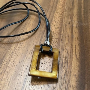 May include: A brown leather cord necklace with a brown rectangular pendant with a smaller rectangular opening in the center. The pendant has a mottled pattern.